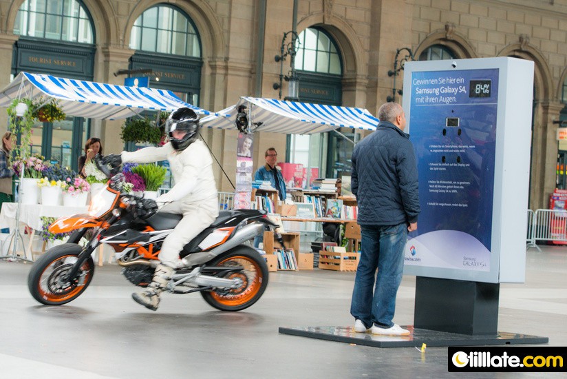 A man stands facing Samsung's All Eyes On The S4 board in a station hall, a phone set into it below a display reading 84%, while a stunt rider on a motorbike rides past