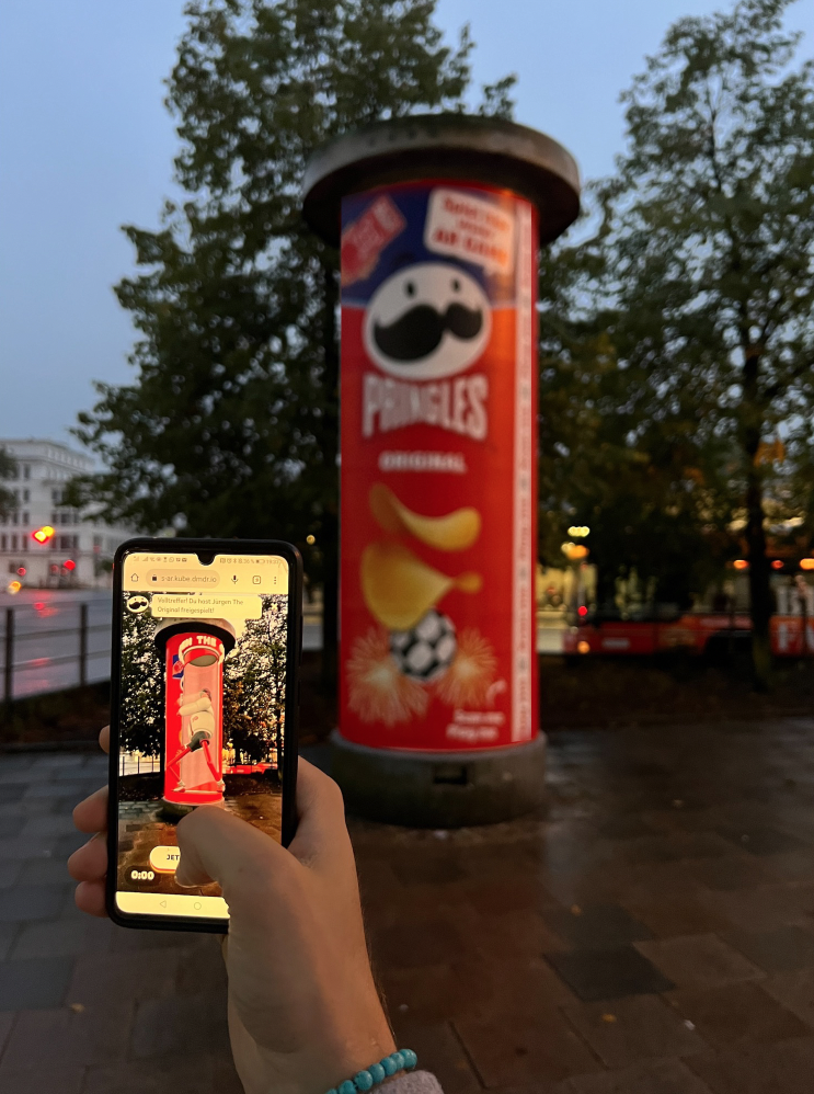 Pringles AR You Ready?!: a phone held up to a street advertising column wrapped as a giant Pringles can; on screen the column's front is open to show a character inside, under a message in German saying Jürgen The Original has been unlocked