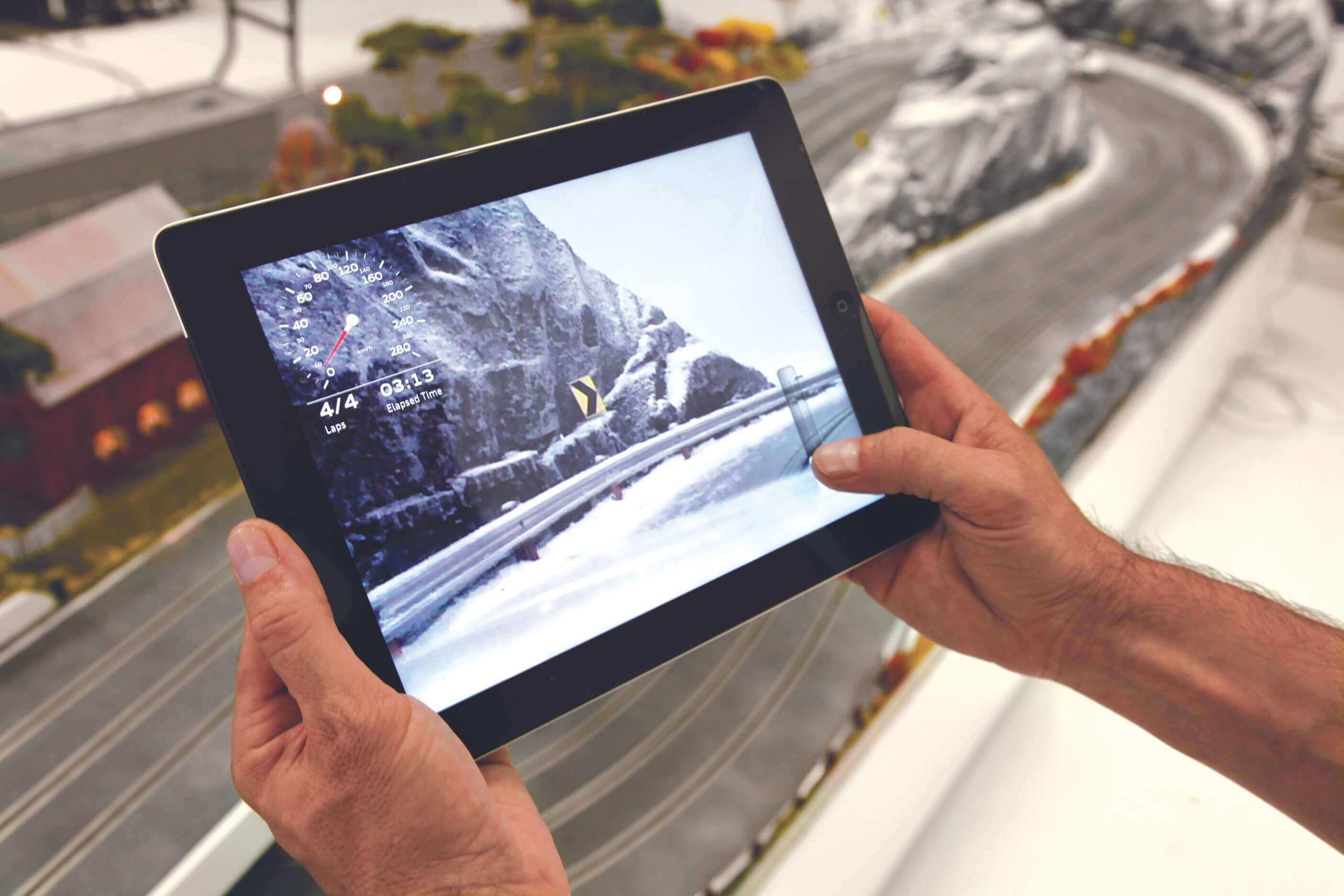 Audi Quattro Experience: hands hold an iPad showing the live in-car view of a snowy mountain pass on the model track, with a speedometer, 4/4 laps and 03:13 elapsed time, the miniature track behind