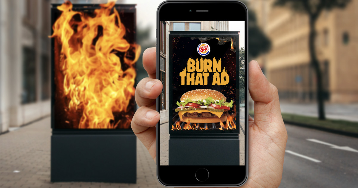 Promotional image for Burn That Ad: a hand holds up a phone whose screen shows a street ad panel as a Burger King poster with a Whopper and flames, beside a panel engulfed in fire