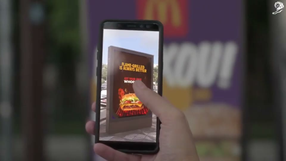 A hand holds a phone up to a McDonald's street poster in Burn That Ad for Burger King; on screen the poster has turned into a Burger King ad reading Flame-grilled is always better, with a free Whopper offer