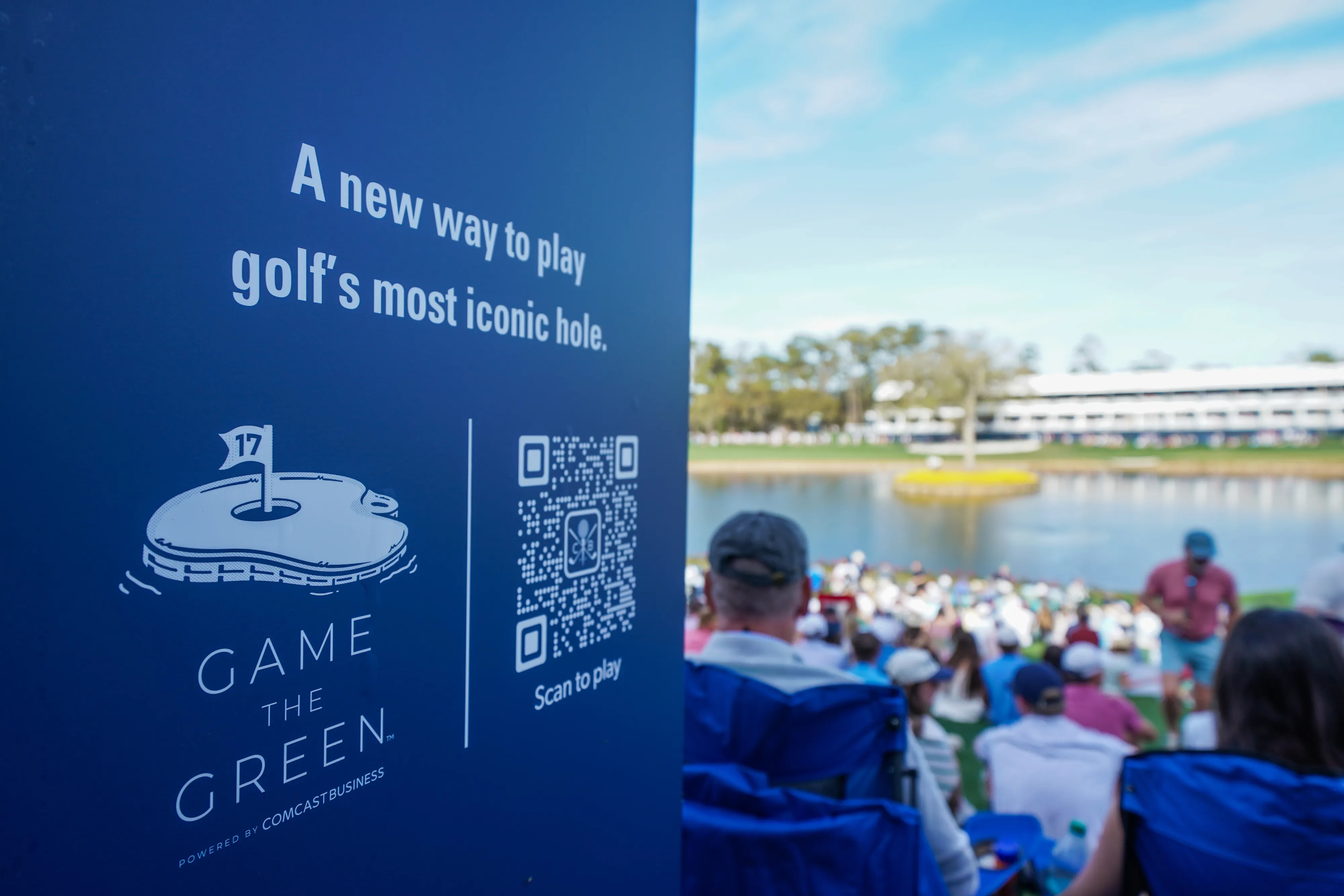 Game The Green sign for Comcast Business with a hole-17 logo and a QR code marked Scan to play, beside spectators in chairs facing an island green across the water