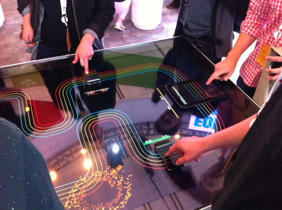 Players at Google's Racer table with fingers on phones and a tablet laid on the glass, coloured race lanes glowing on the display beneath