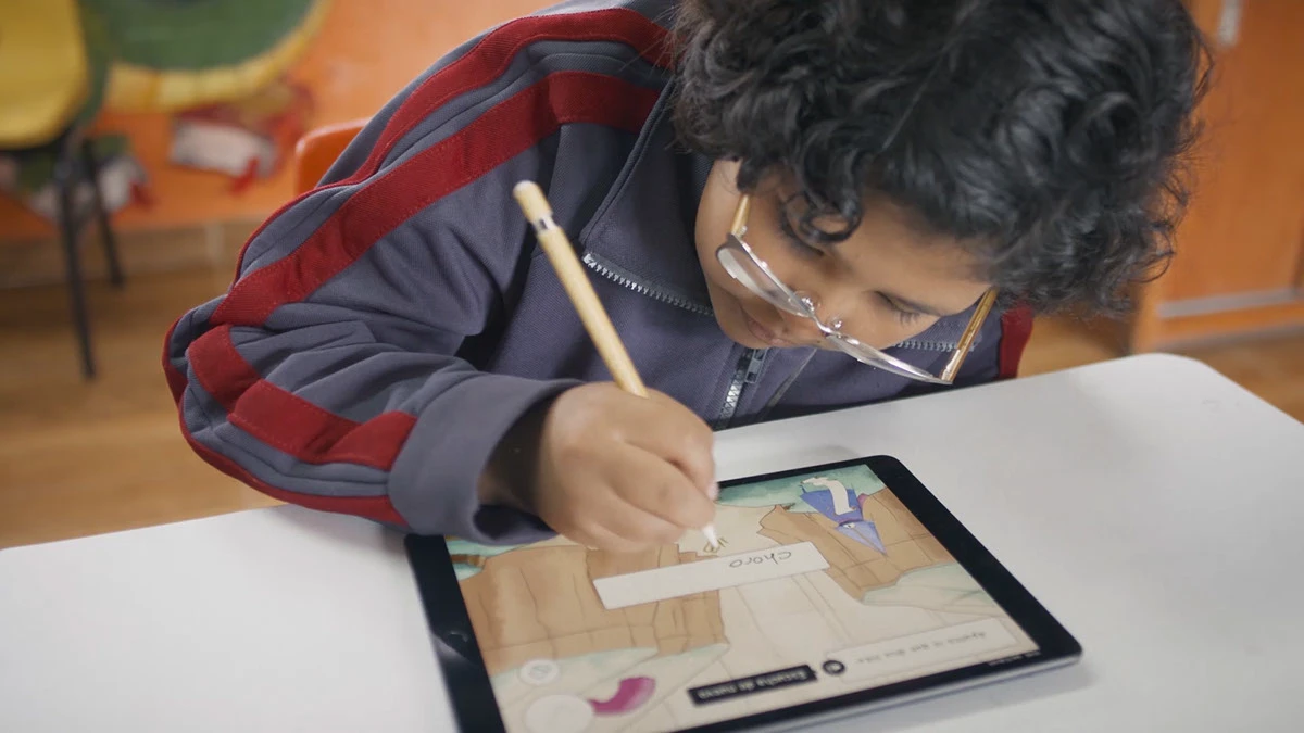 A child writes a word with a stylus on a tablet showing the Kiddi World game for Pharmaton