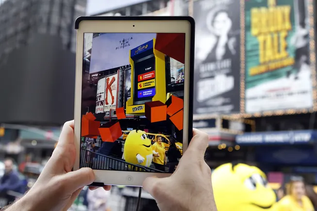 Hands hold up a tablet among big theatre billboards; on screen a yellow M&M's arcade cabinet in augmented reality offers Caramel Cannon, Caramel Crawl and Square Smash, with orange cubes floating beside a costumed yellow M&M's character