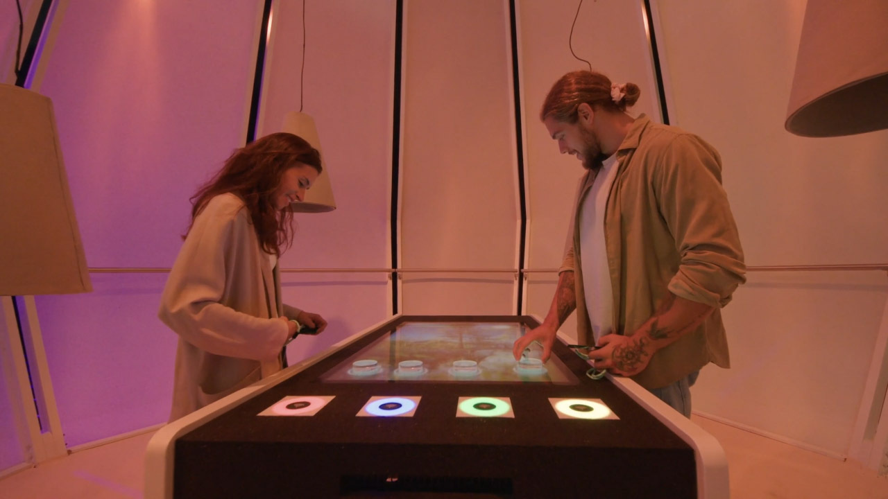 Skoda Make Every Km Count: a woman and a man stand at a touch table in a pink-lit pod, the man's hand on one of four round tokens resting on an outdoor scene shown on the screen, with four glowing coloured rings along the table's front edge