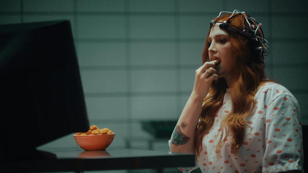 A woman in an electrode headset eats a snack from a bowl beside a screen, in Seara's Mind Player