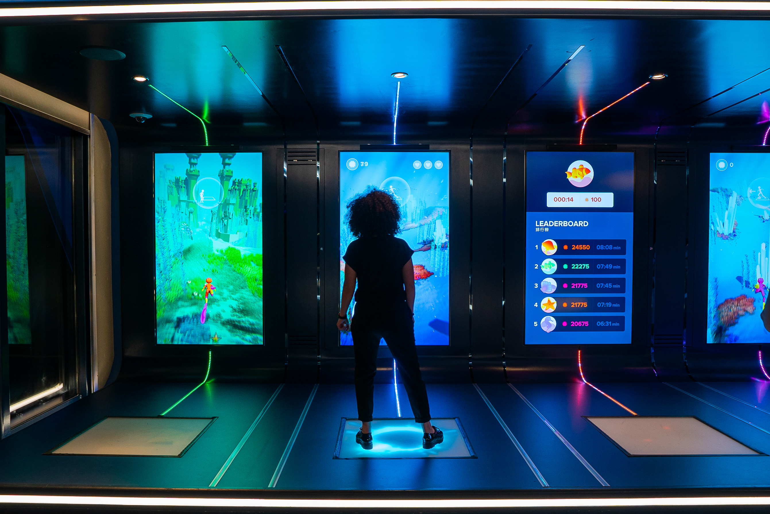 Sasu's Mystical Quest for Royal Caribbean: a player stands on a glowing floor board facing a tall screen with an underwater level, between other game screens and a top-five leaderboard