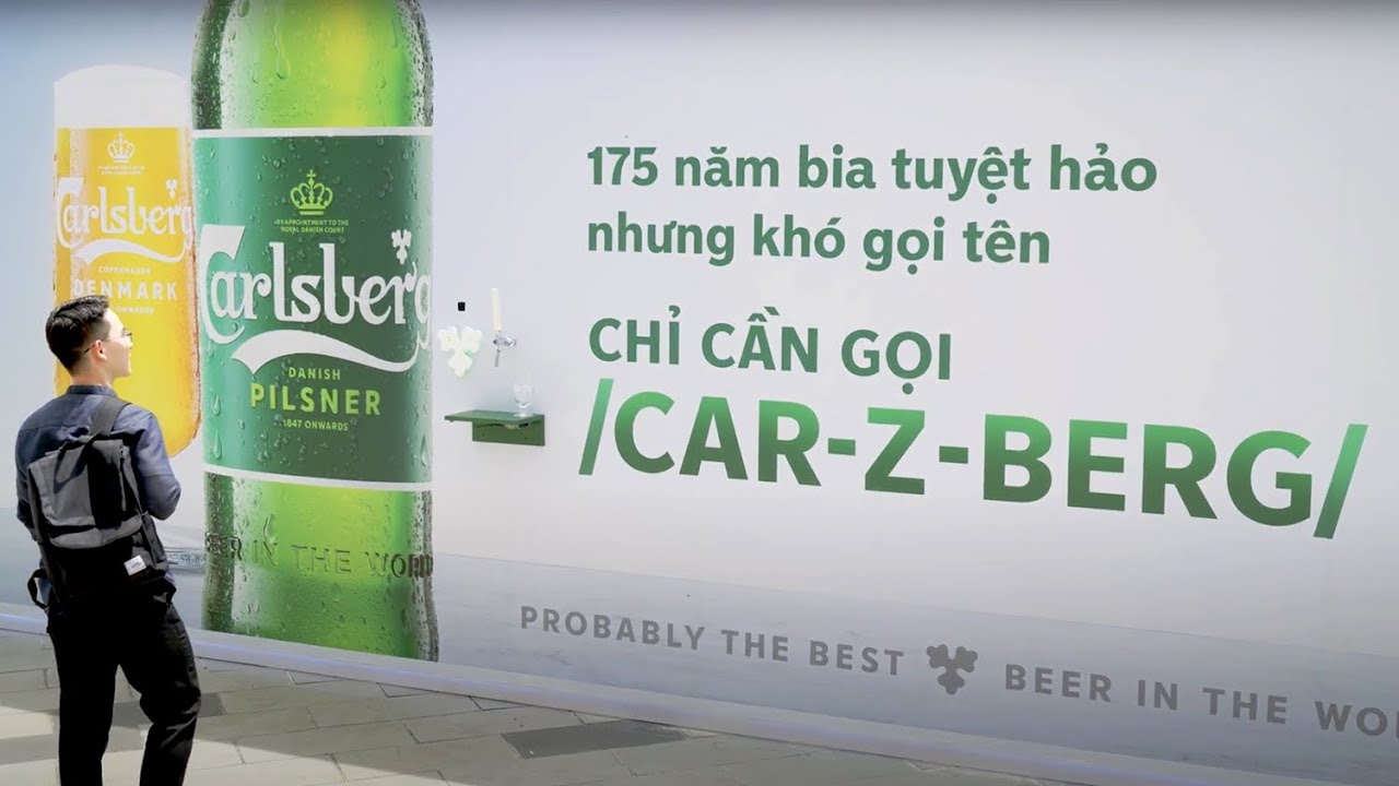 A young man with a backpack stands before Carlsberg's Say Carlsberg billboard, which shows a Danish Pilsner bottle, a beer tap above a small green shelf and the phonetic cue /CAR-Z-BERG/ under Vietnamese text