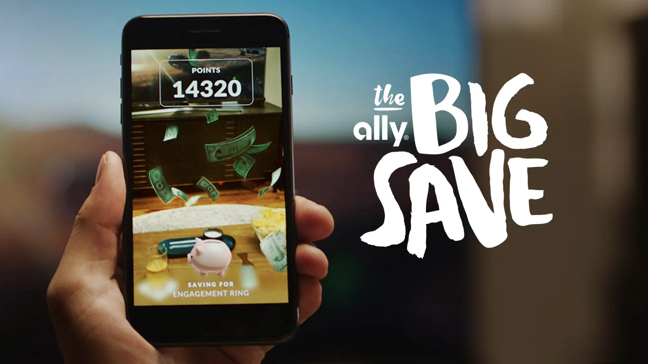 A hand holds a phone running Ally Bank's The Big Save: green dollar bills fall over a living room towards a pink piggy bank on the coffee table, with 14320 points at the top and Saving for engagement ring below, beside the game's logo