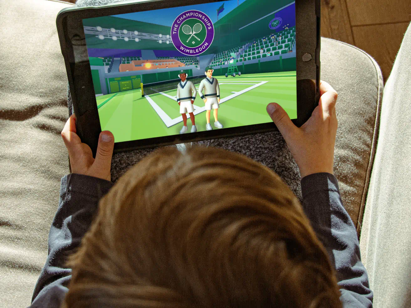 Seen over a child's head, a tablet shows WimbleWorld on Roblox: two avatars in cream V-neck tennis sweaters stand on a grass court under the Wimbledon Championships crest, with green stands behind